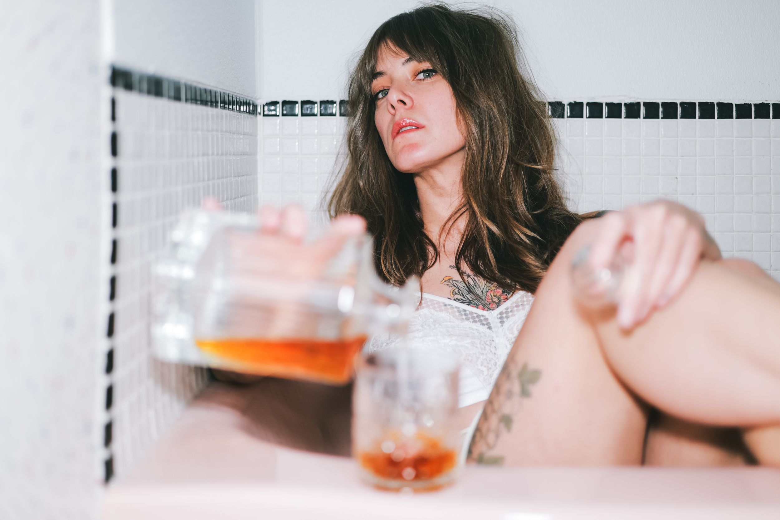 Audrey Martin in white lace sits in a tiled bathtub, pouring whisky into a glass toward the camera, Calgary