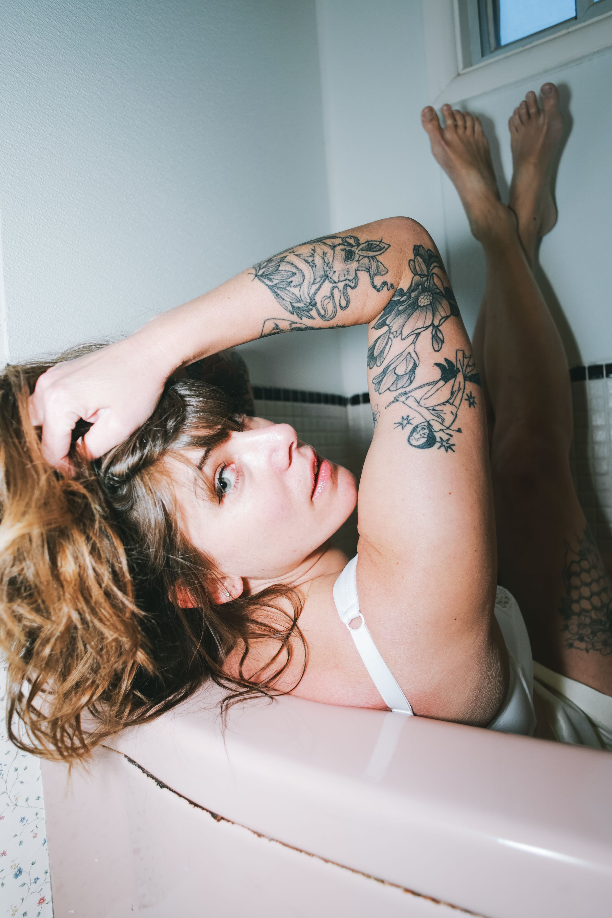 Audrey Martin, sleeve tattoos showing, rests her head on a pink bathtub edge with legs raised on the tiled wall, Calgary