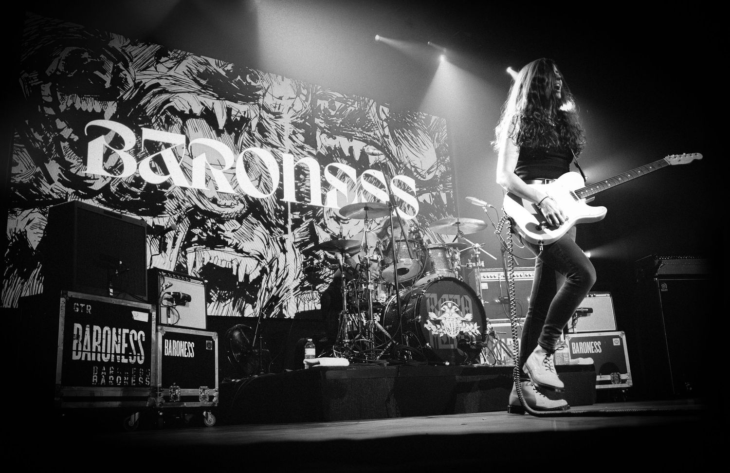 Long-haired Baroness guitarist at stage right beside the drum kit and illustrated logo backdrop, The Palace Theatre, Calgary