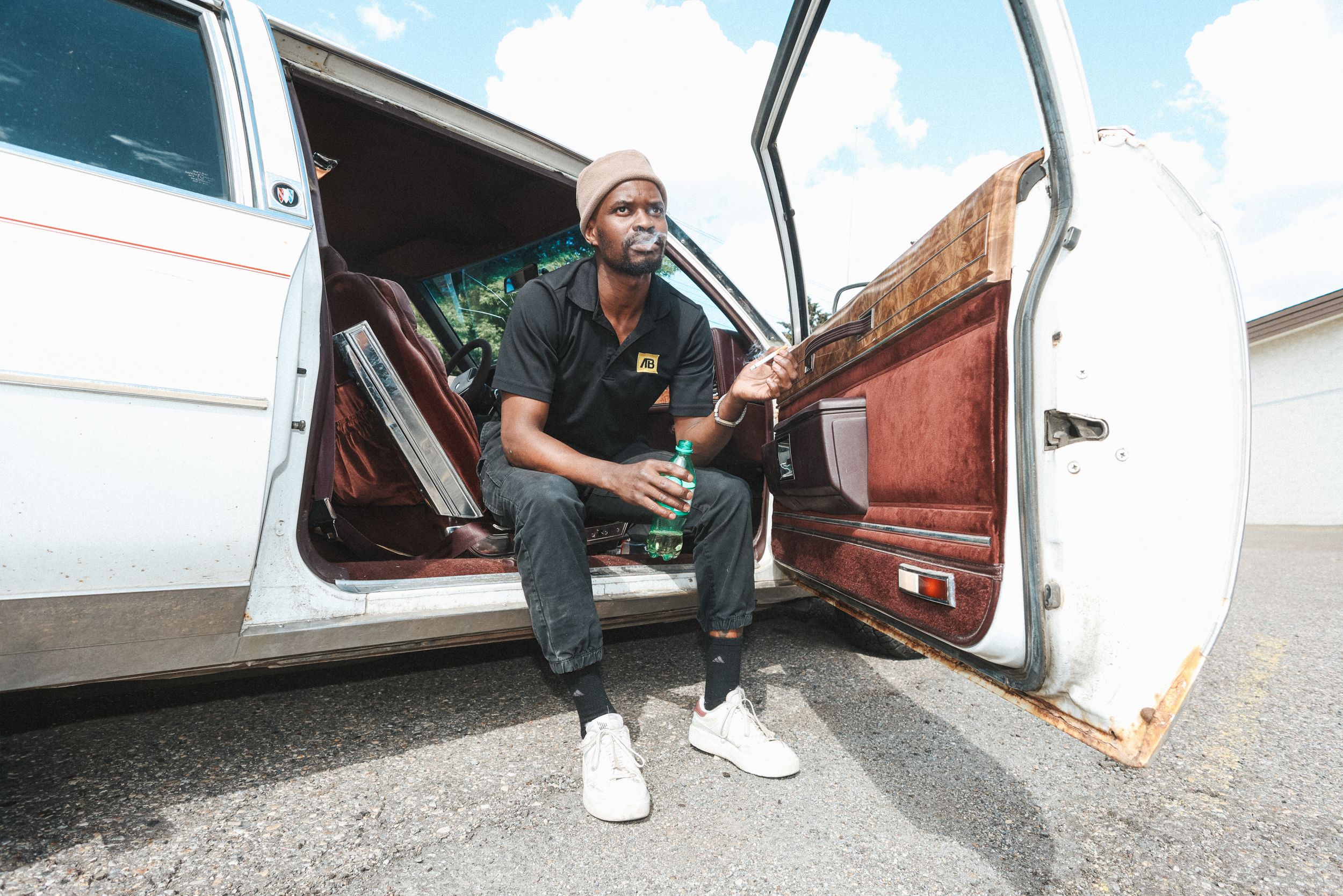 Man in a beanie and black polo sits in the open door of a white vintage car with red seats, holding a green bottle, Calgary
