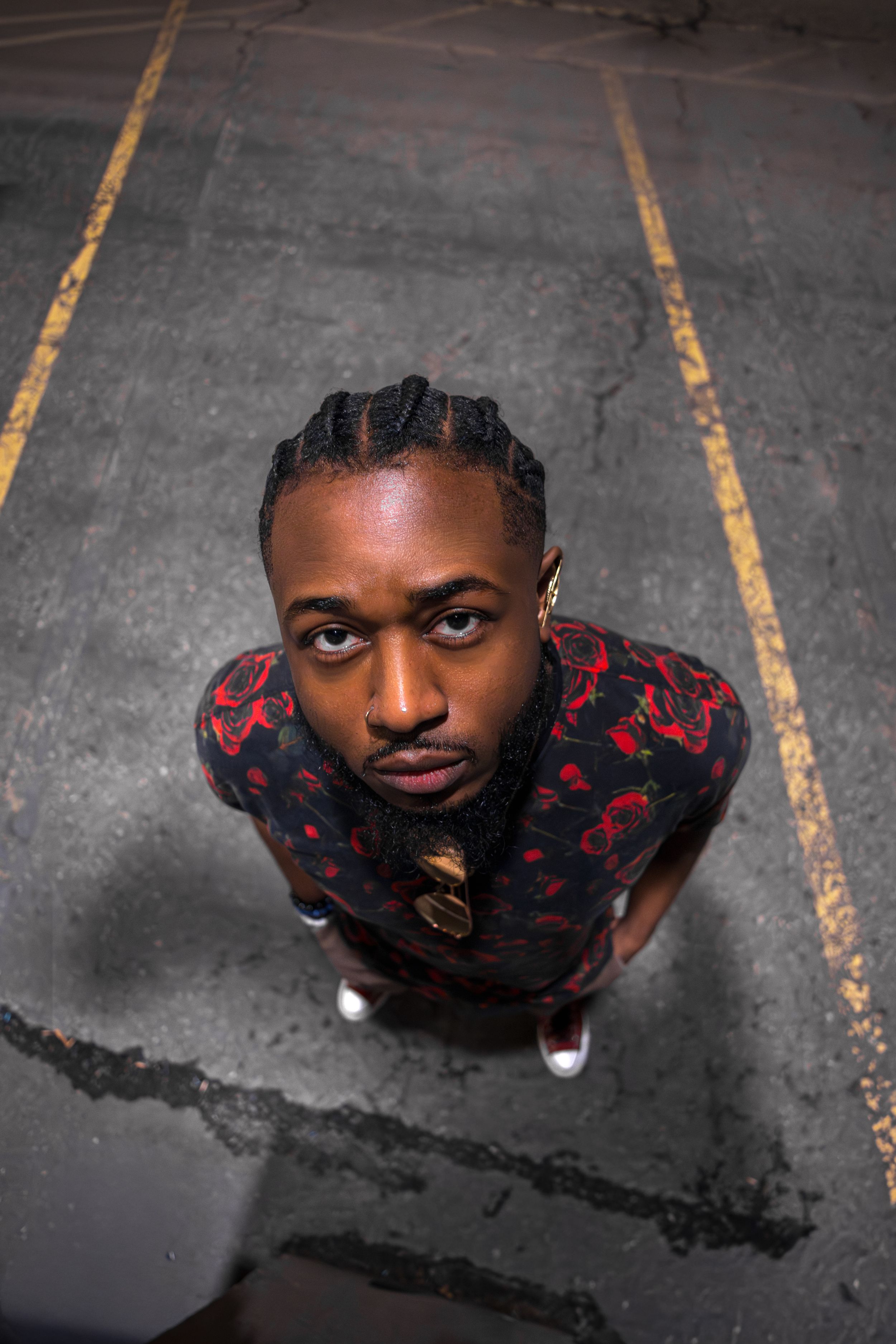 Overhead wide-angle portrait of a man with braids in a red floral print shirt, looking up from a Calgary parking lot