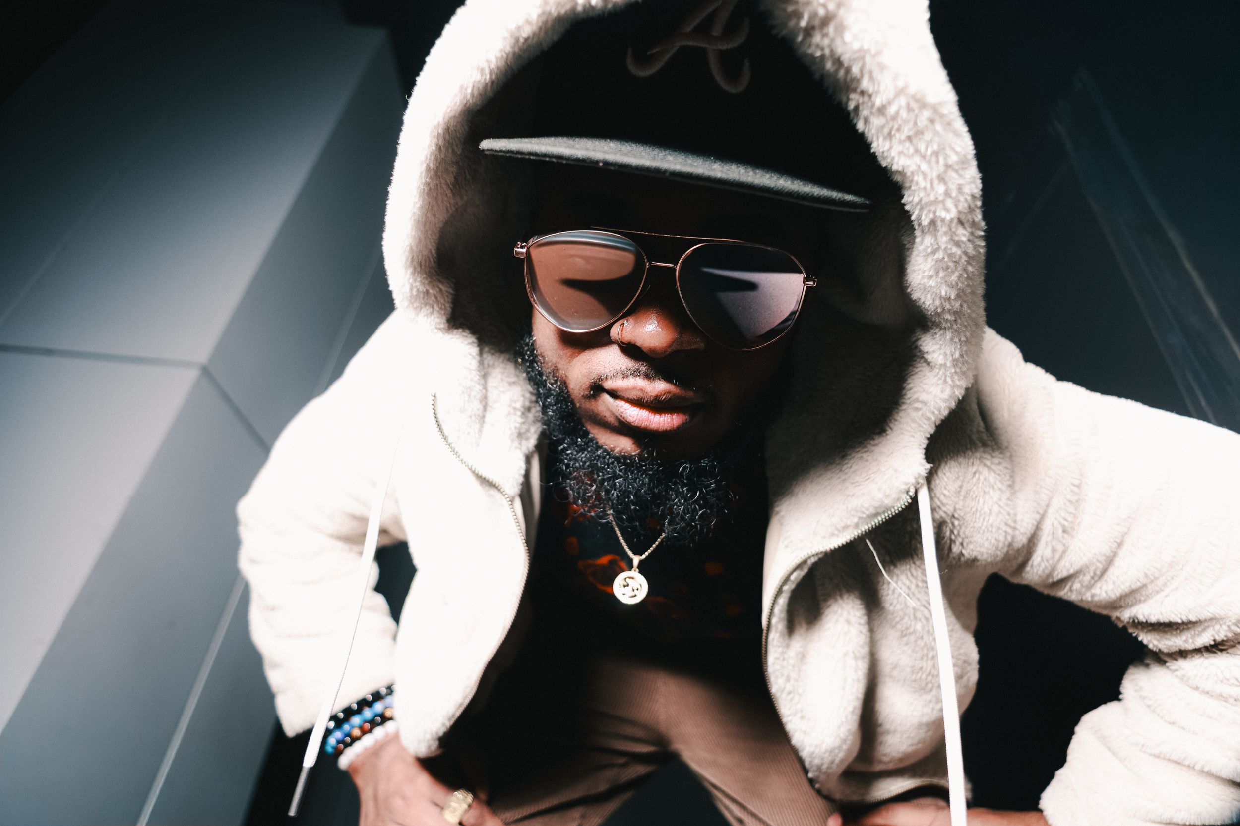 Man in a white sherpa hoodie, cap and aviator sunglasses crouches toward the lens, gold pendant catching light, Calgary