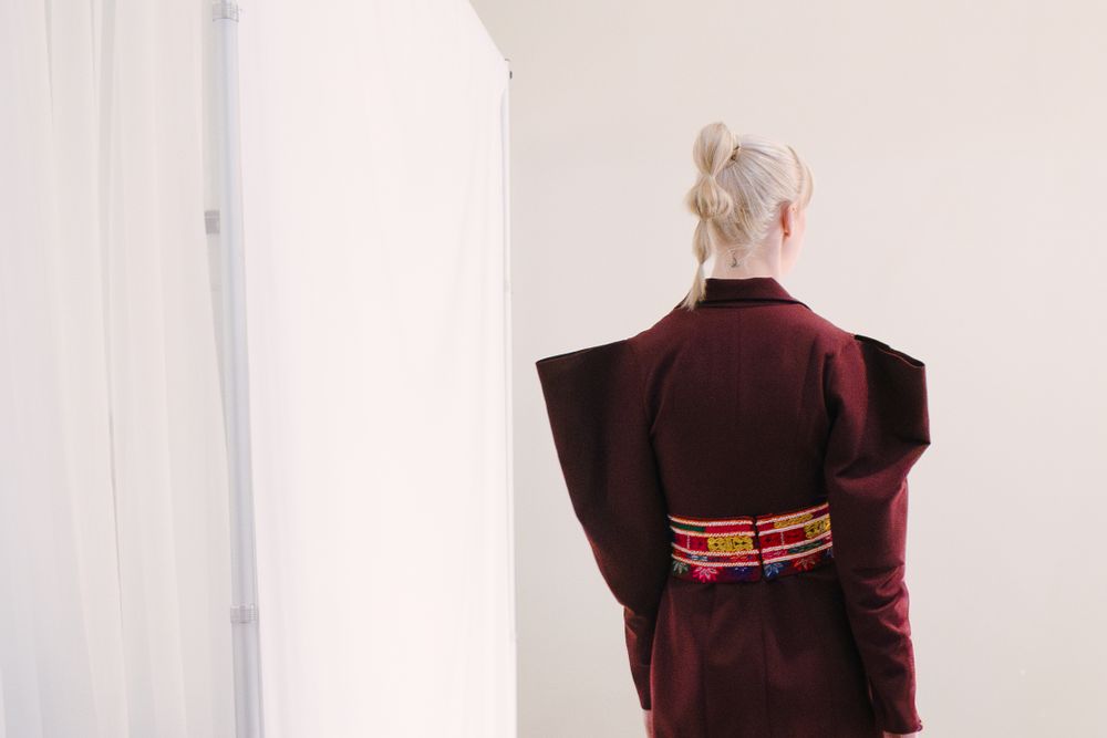 Back view of a platinum blonde model in a burgundy jacket with sculpted shoulders and a woven belt, Edmonton Fashion Week