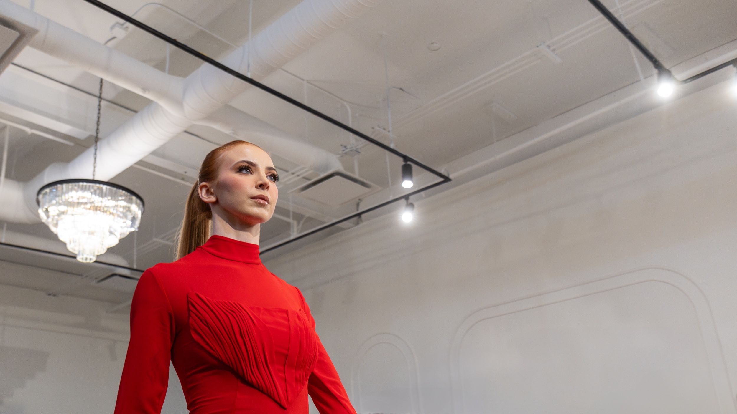 Model in a fitted red gown walks beneath a crystal chandelier in a white gallery space, Edmonton Fashion Week 2024
