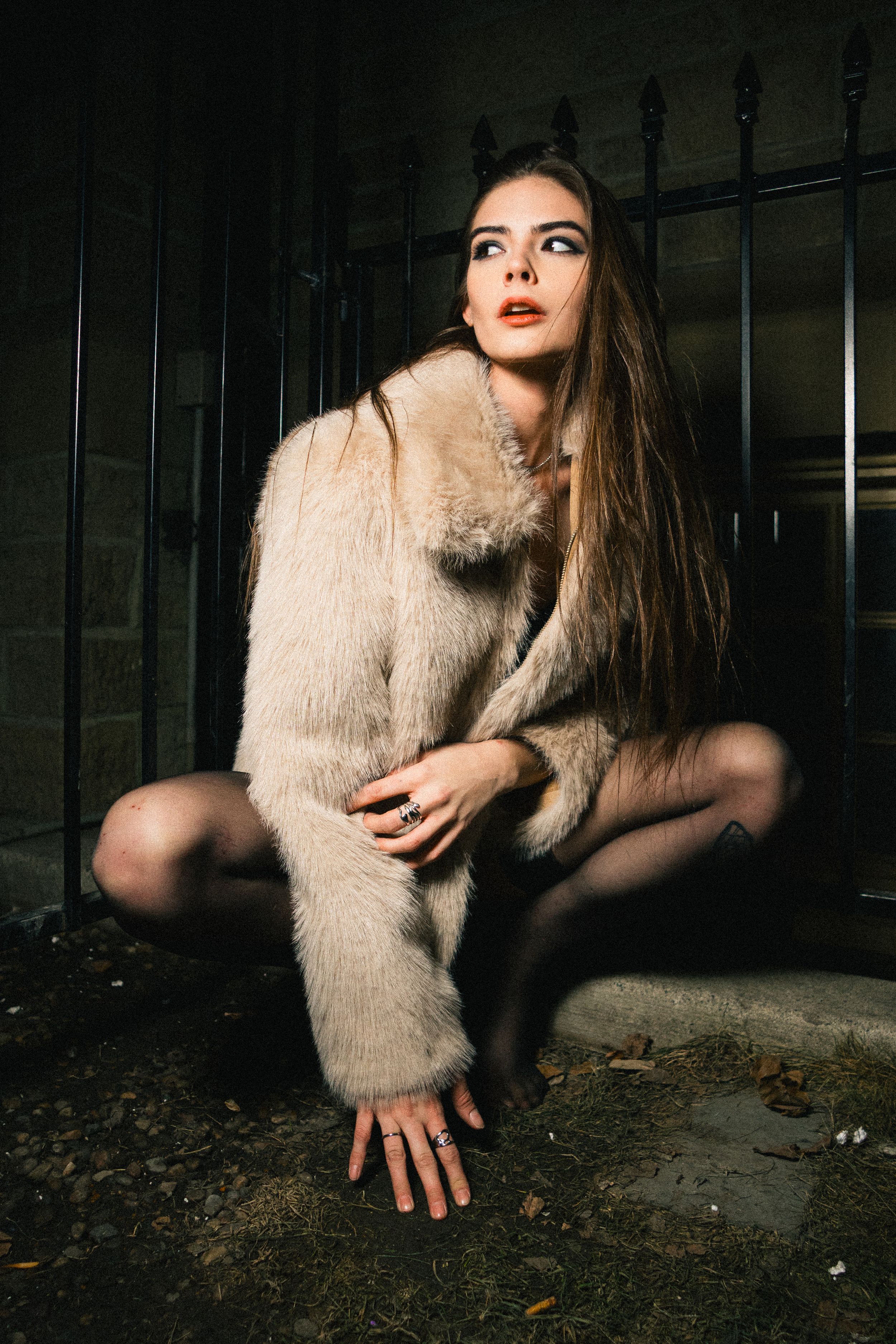 Model in a cream faux fur coat crouches before a black iron gate at night, hand on the ground, Gutter Glam series, Calgary