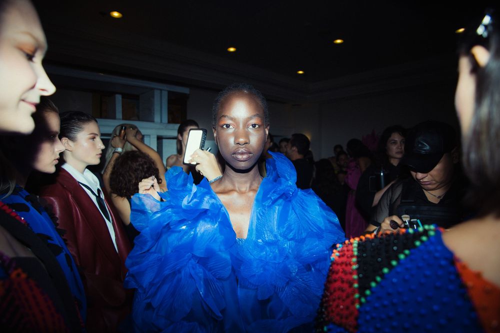 Backstage at Mexico City Fashion Week, a model in a vivid blue ruffled tulle gown stands among models and crew with phones