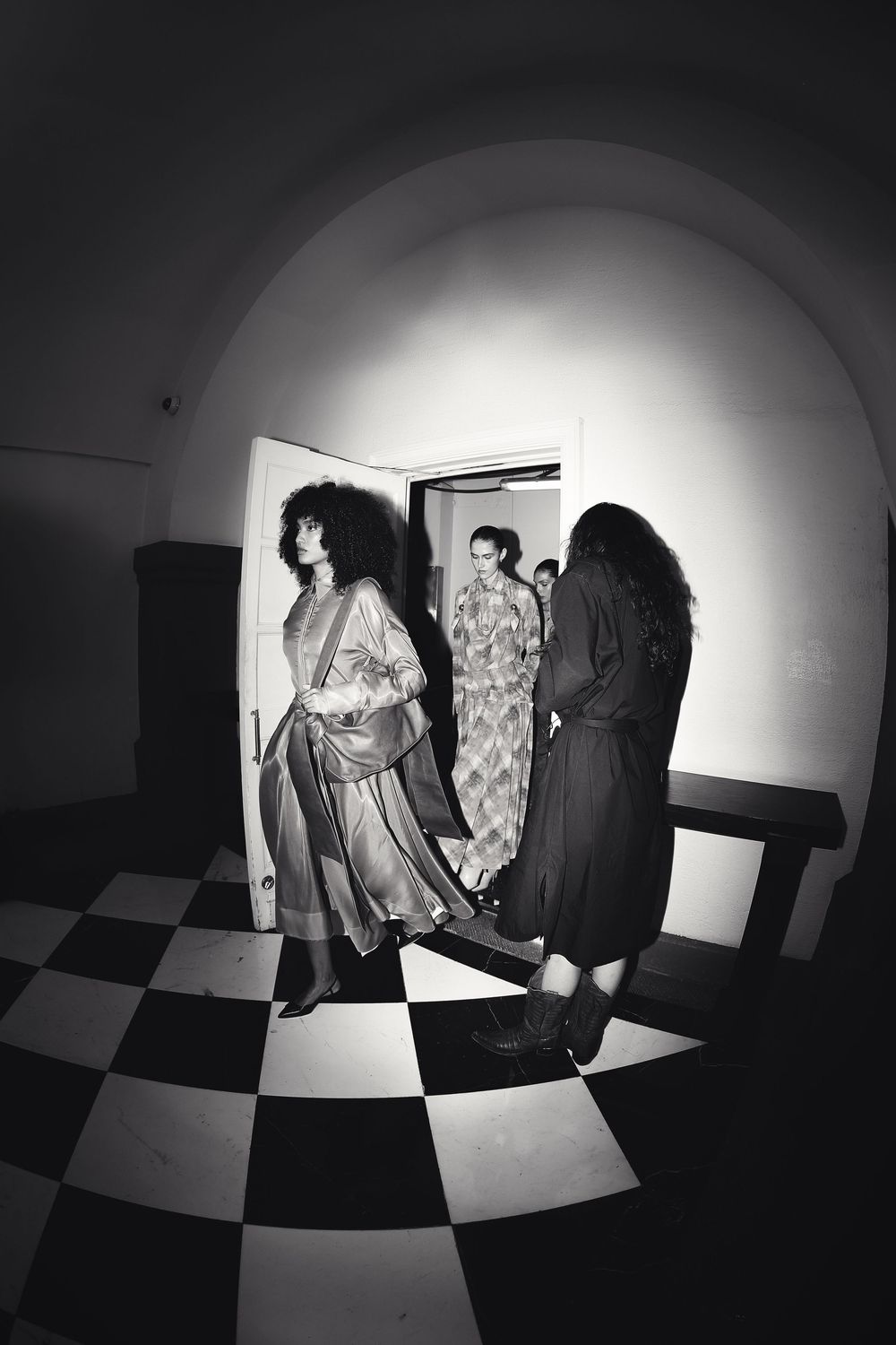 Black and white image of models passing a doorway onto a checkered floor backstage at Mexico City Fashion Week