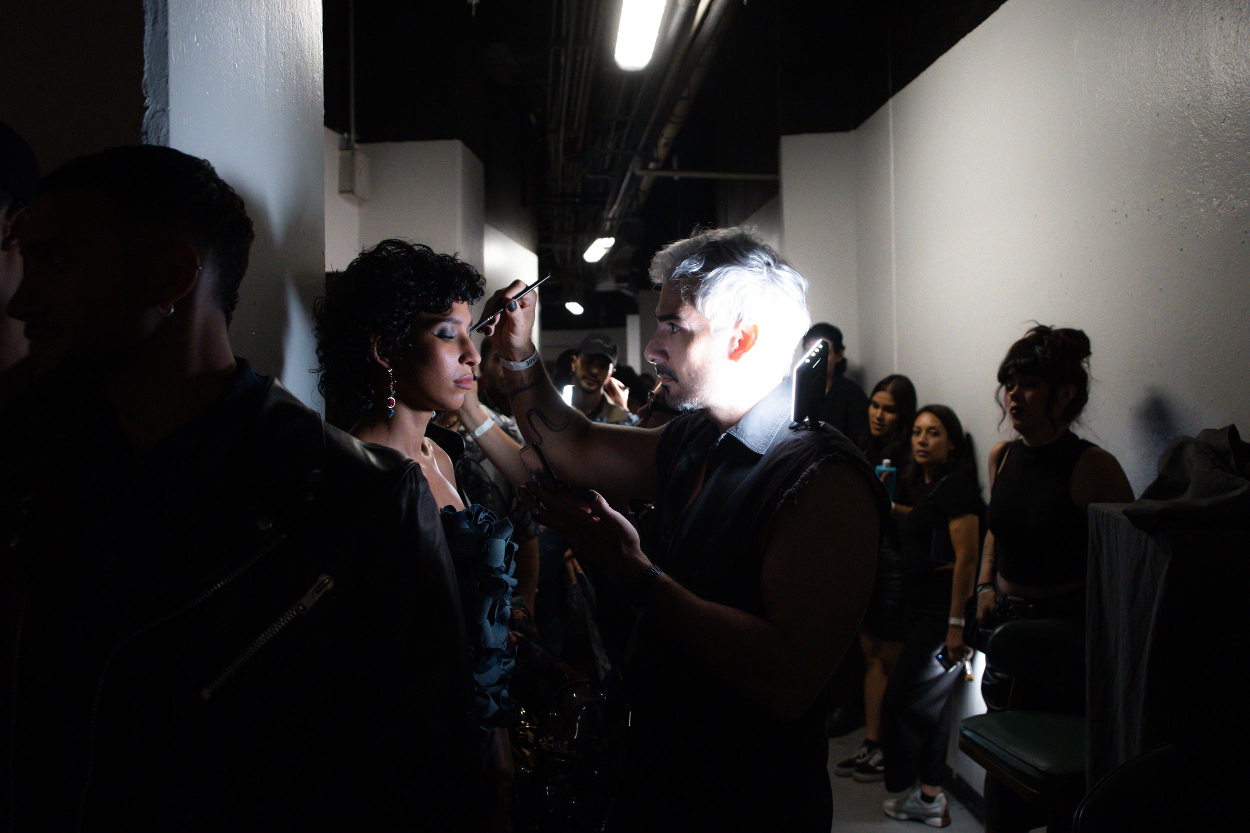 Makeup artist prepares a model with dark curly hair in a dim backstage corridor at Mexico City Fashion Week
