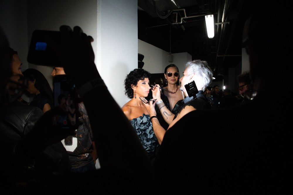 Backstage at Mexico City Fashion Week, a makeup artist touches up a model in a blue sequin top under hard light