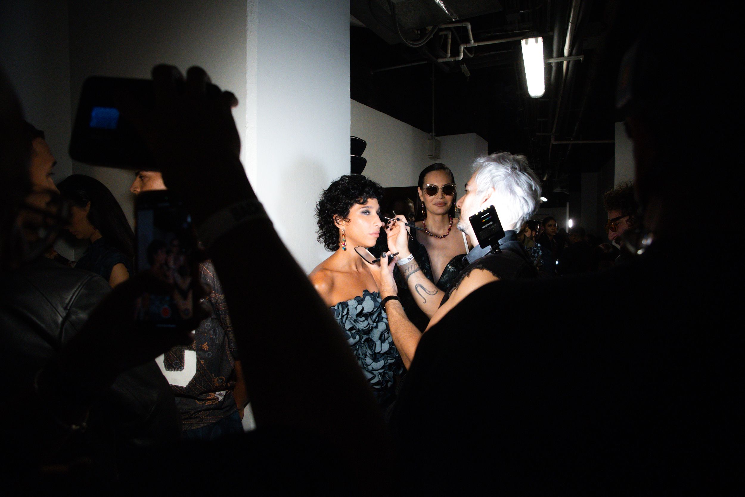 Makeup artist works on a model in a blue sequined dress backstage at Mexico City Fashion Week, a camera in the foreground