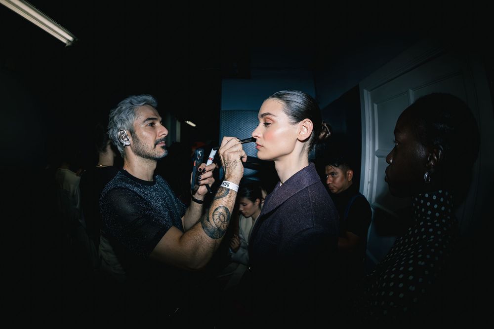 Tattooed makeup artist applies final touches to a model in a burgundy blazer backstage at Mexico City Fashion Week