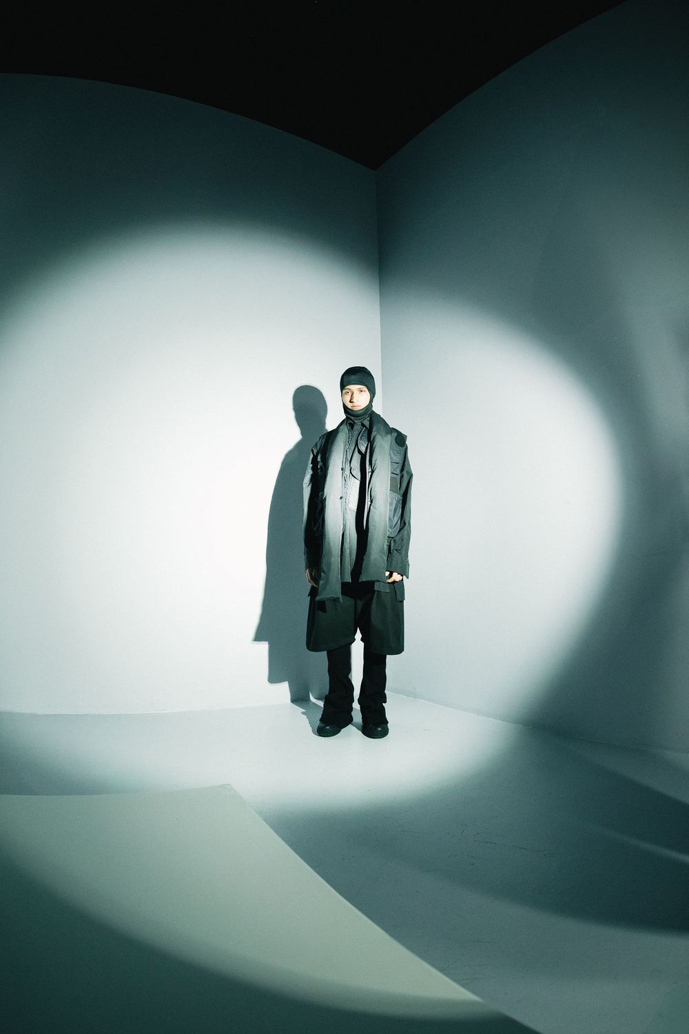 Model in a dark layered coat and balaclava in a spotlit white corner, casting a long shadow, at Mexico City Fashion Week