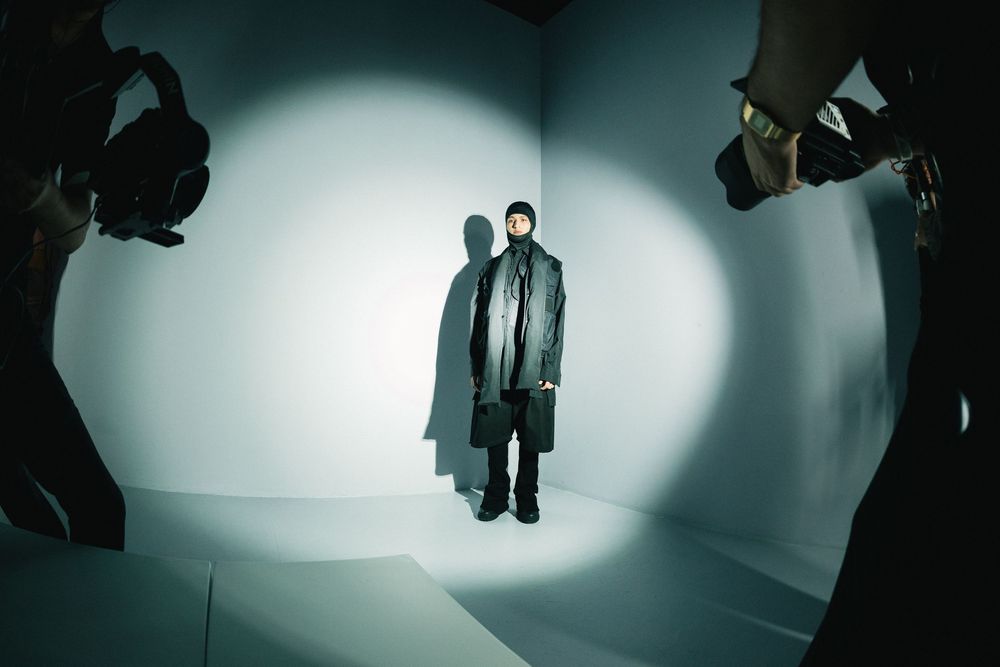 Model in a long black coat and balaclava in a spotlight on a white set, framed by cameras, Mexico City Fashion Week
