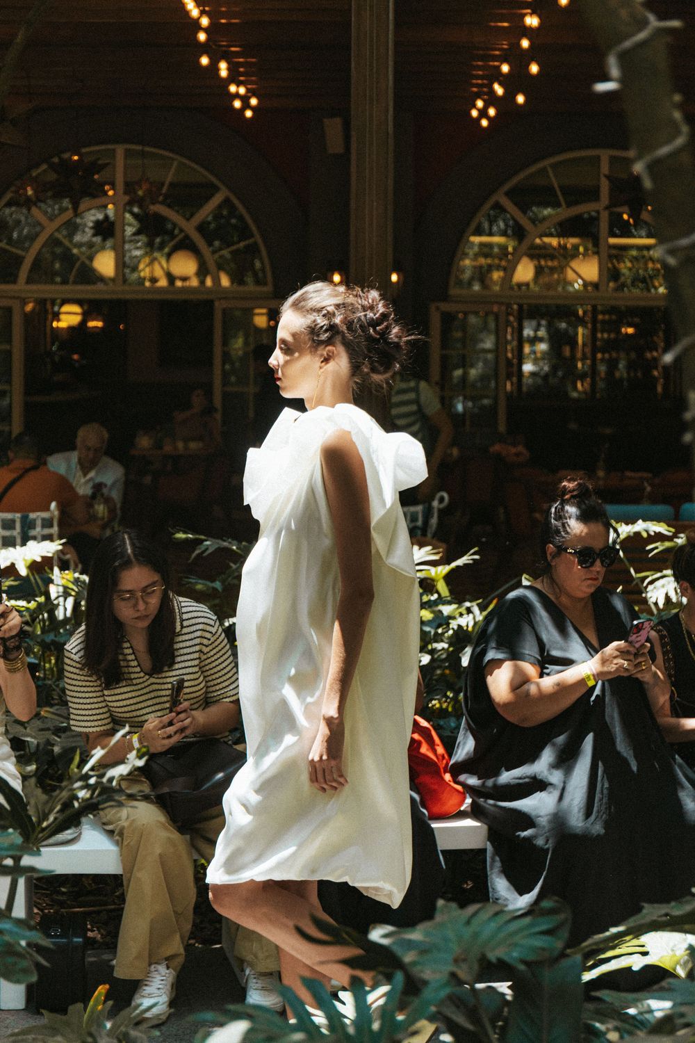 Model in a white draped cape dress walks a sunlit garden runway under arched windows at Mexico City Fashion Week