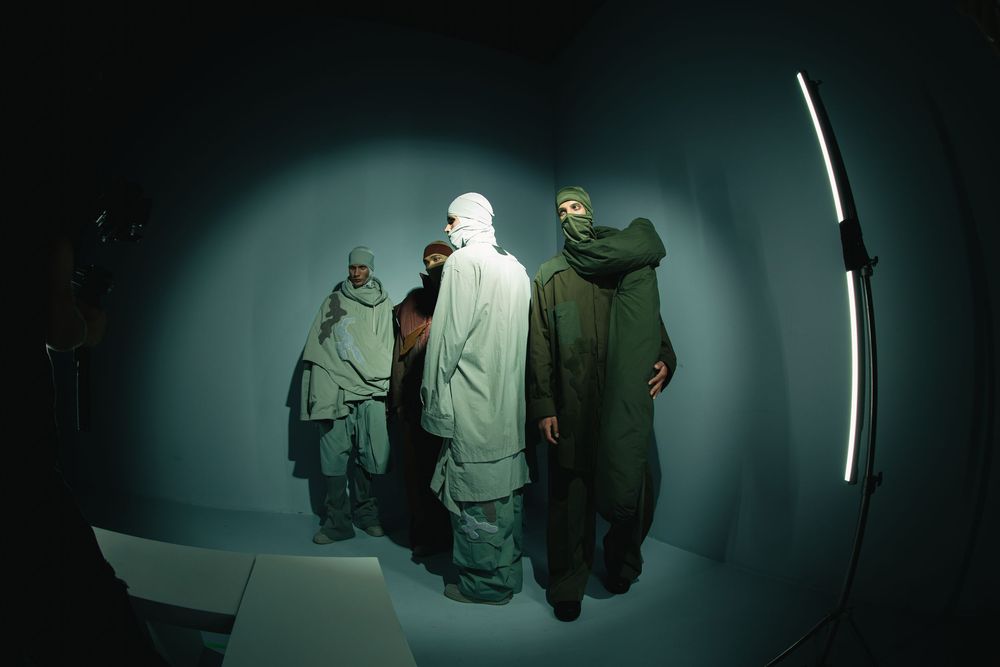 Four models in hooded olive and sage layers with face coverings in a teal-lit studio, fisheye view, Mexico City Fashion Week