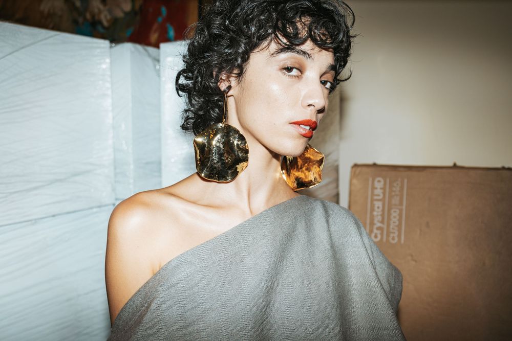 Curly-haired model in a grey off-shoulder top with large gold sculptural earrings and red lipstick, Mexico City Fashion Week