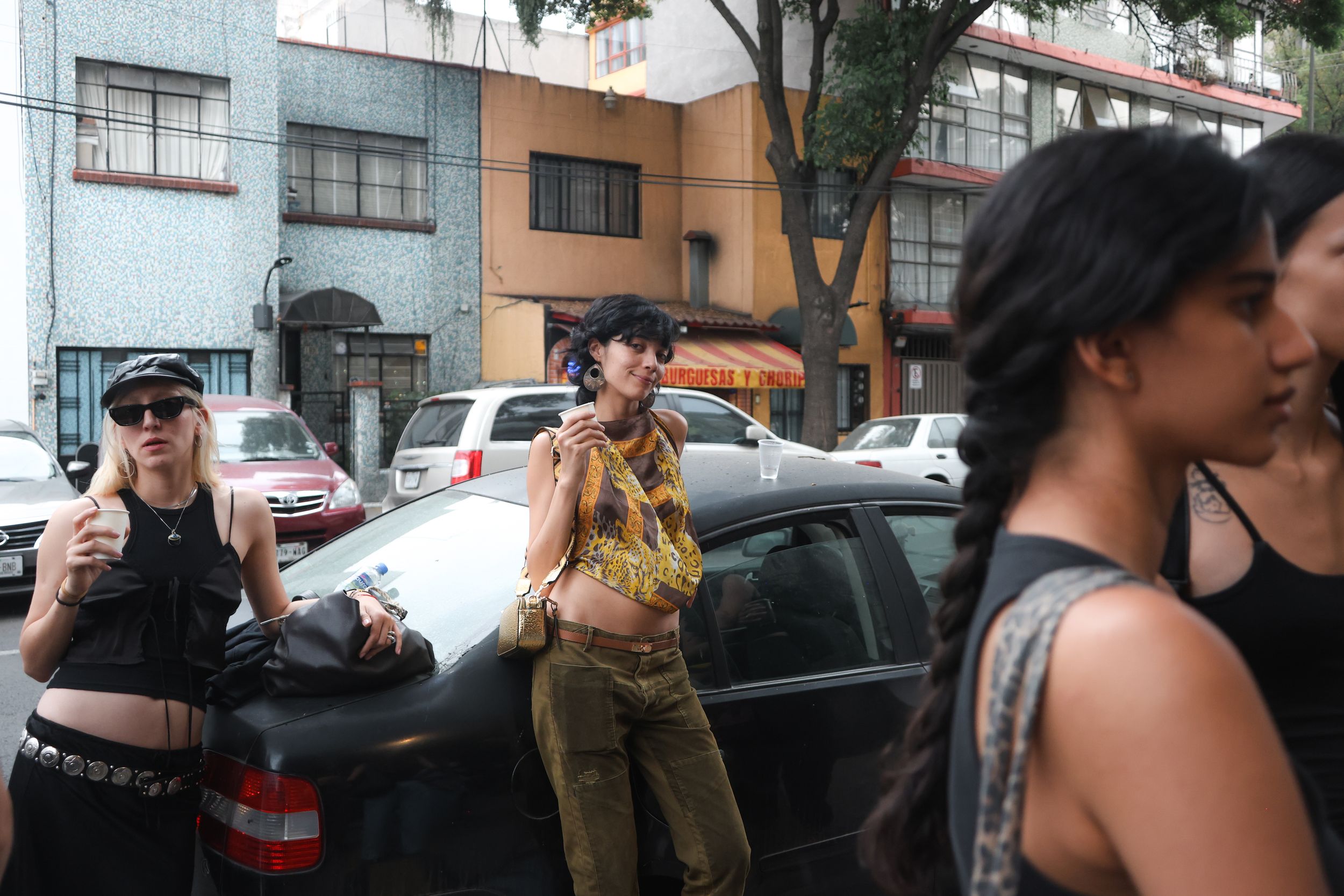 Models wait on a street between shows at Mexico City Fashion Week, one in a gold halter top leaning on a black car