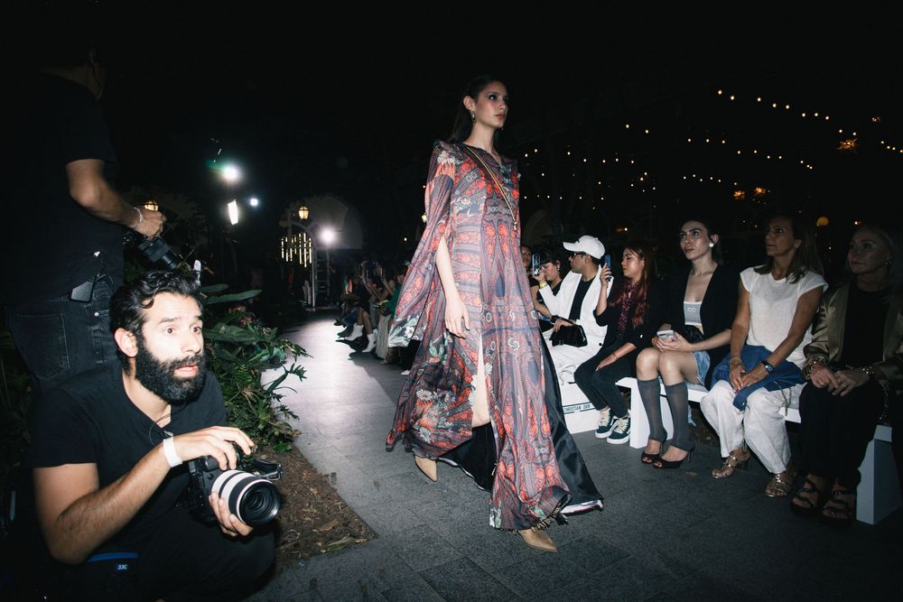 Model in a flowing printed pink and red gown walks a night runway as a photographer crouches, Mexico City Fashion Week