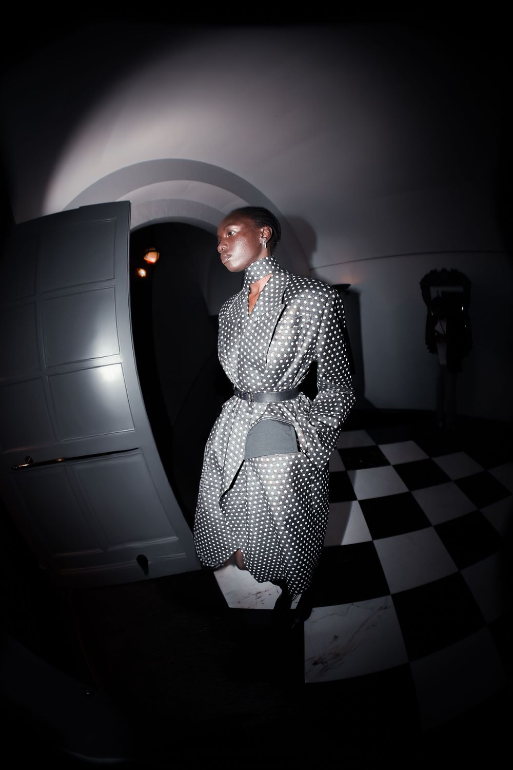 Black and white fisheye image of a model in a belted polka dot coat on a checkerboard floor, Mexico City Fashion Week