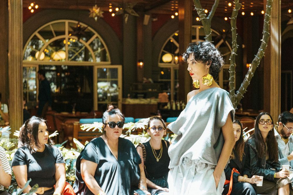 Model in a grey draped top and gold collar walks a courtyard runway under string lights at Mexico City Fashion Week
