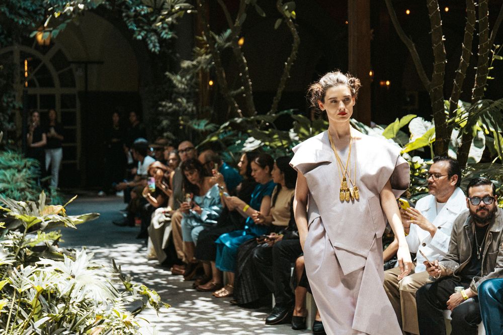 Model walks a garden courtyard runway in a lilac draped dress and gold pendant at Mexico City Fashion Week, among plants
