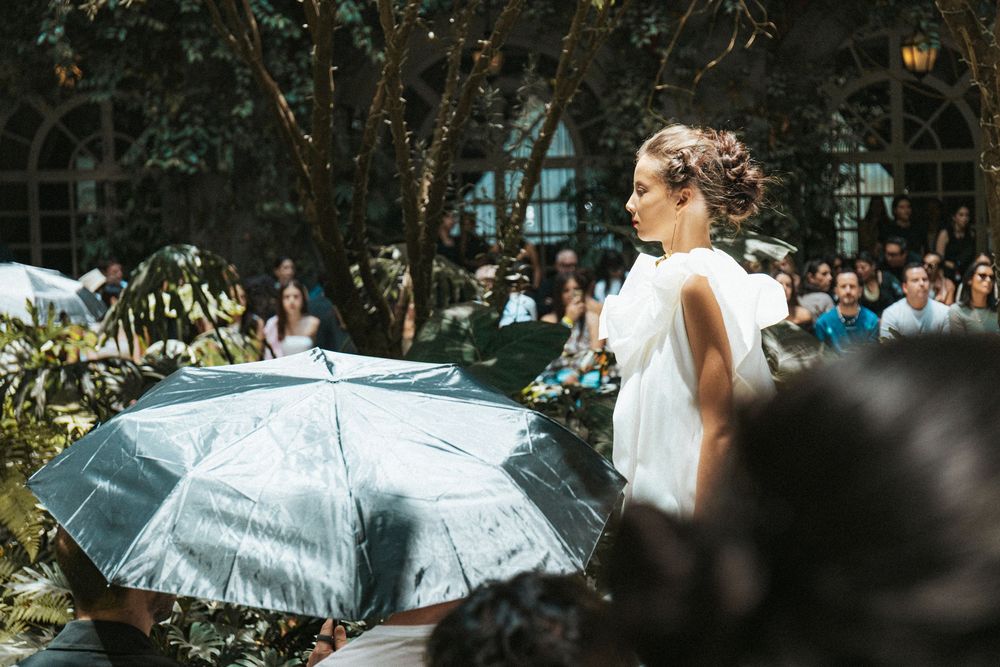 Model with braided updo in a white draped gown walks a leafy courtyard runway at Mexico City Fashion Week