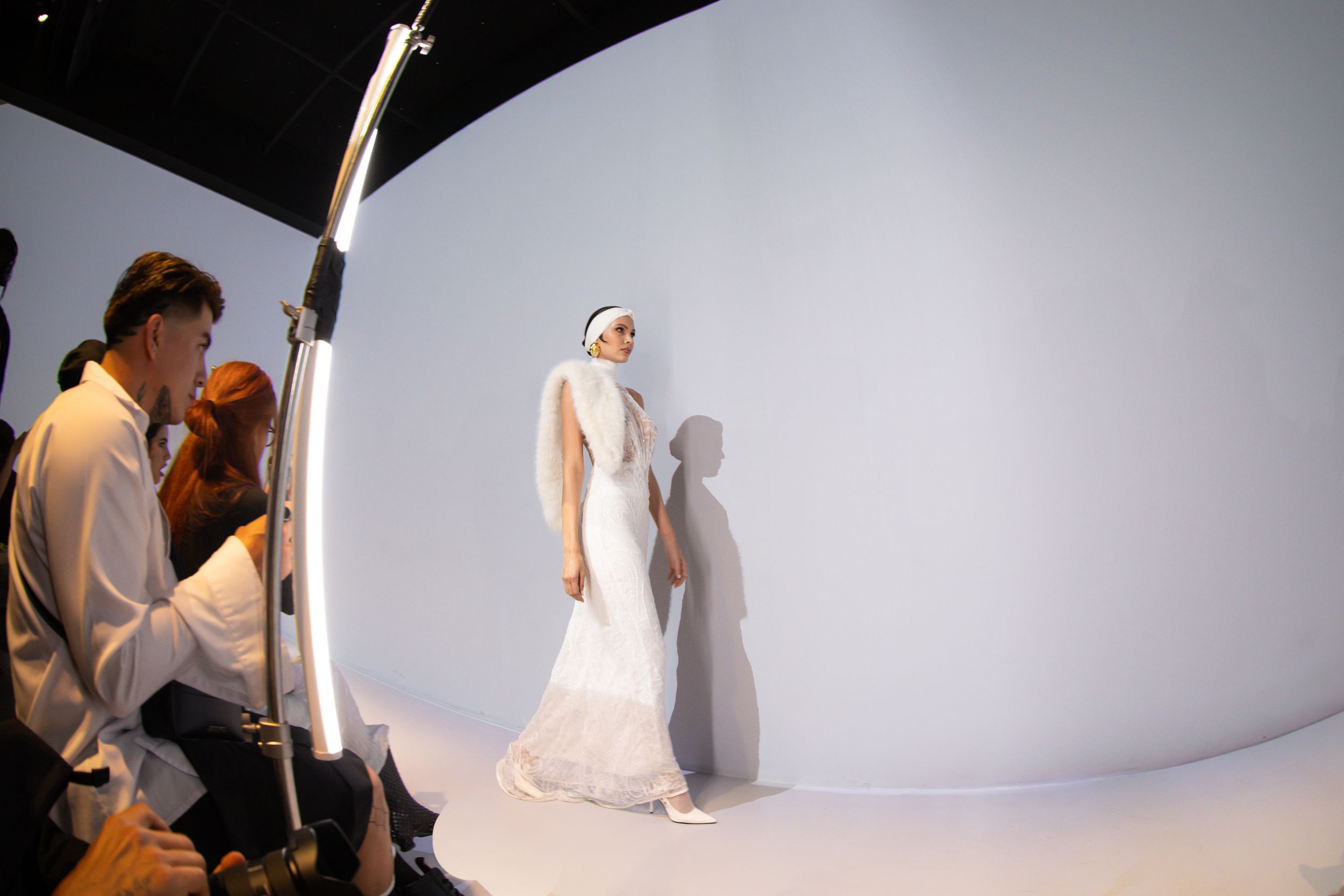 Model in a white lace gown and shawl walks a curved white set past a ring light and photographers, Mexico City Fashion Week