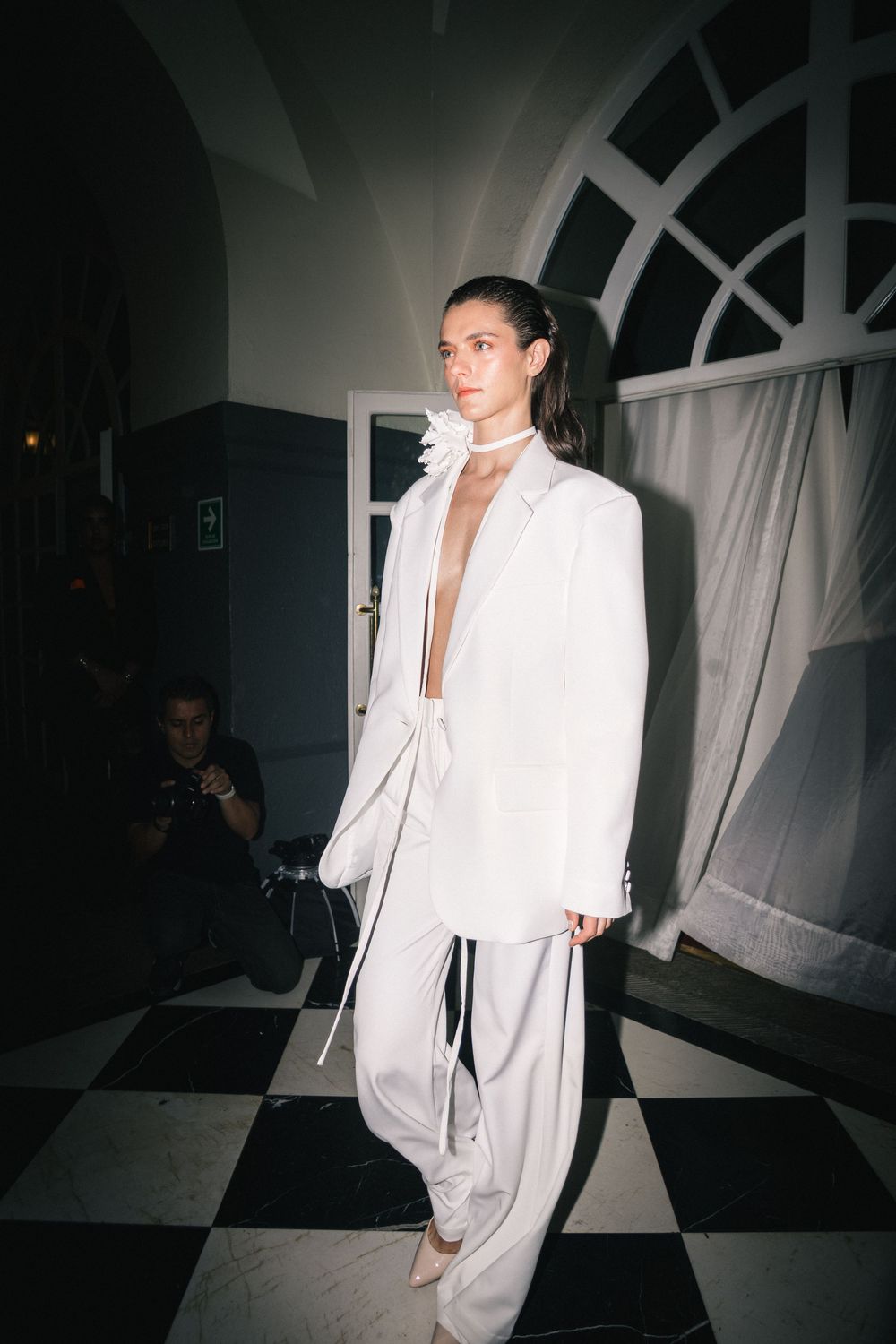 Model in a white oversized suit with plunging blazer and rosette choker walks a checkerboard floor, Mexico City Fashion Week