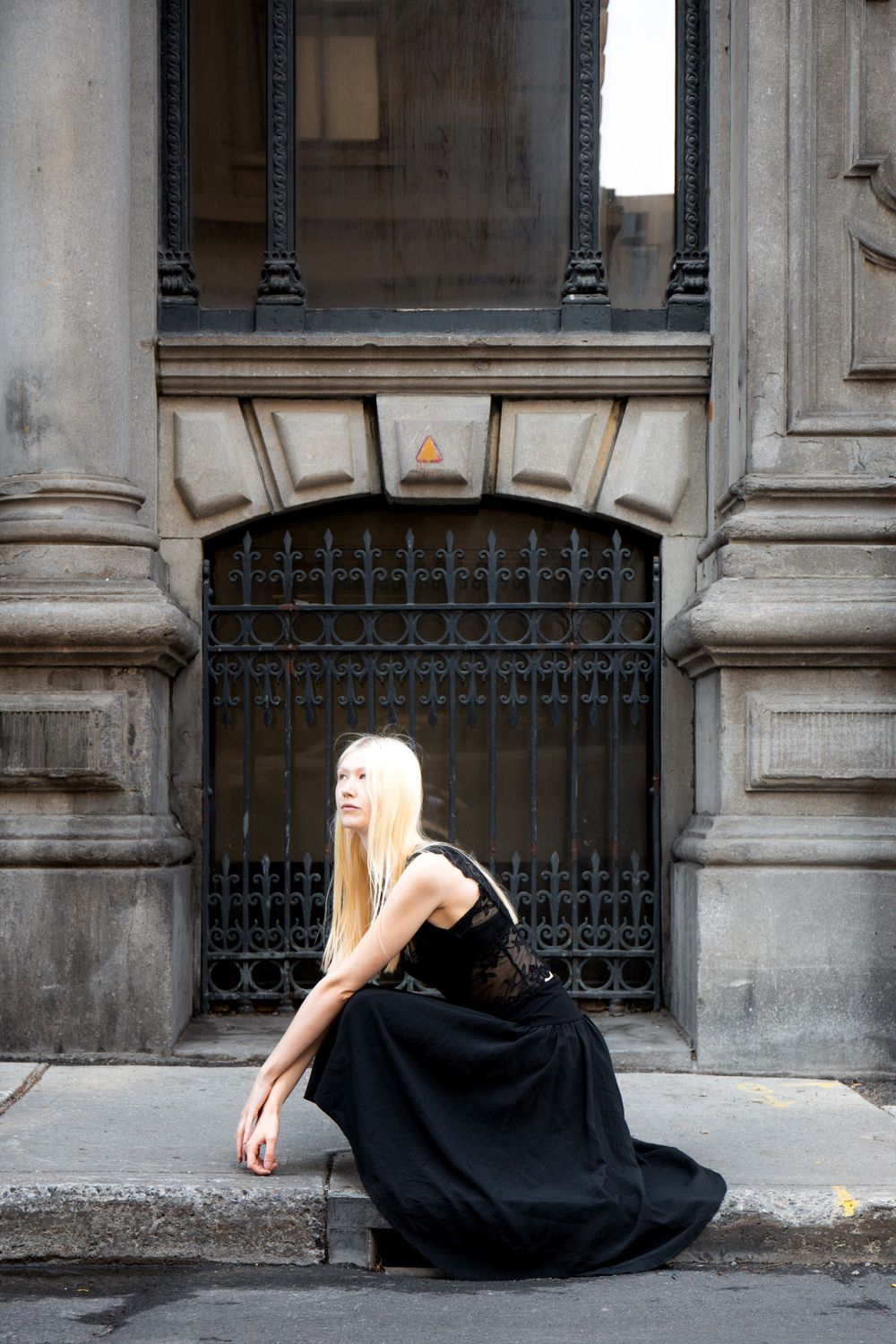 Long-haired blonde model in a black lace gown kneeling before an iron-gated stone doorway during Montreal Fashion Week