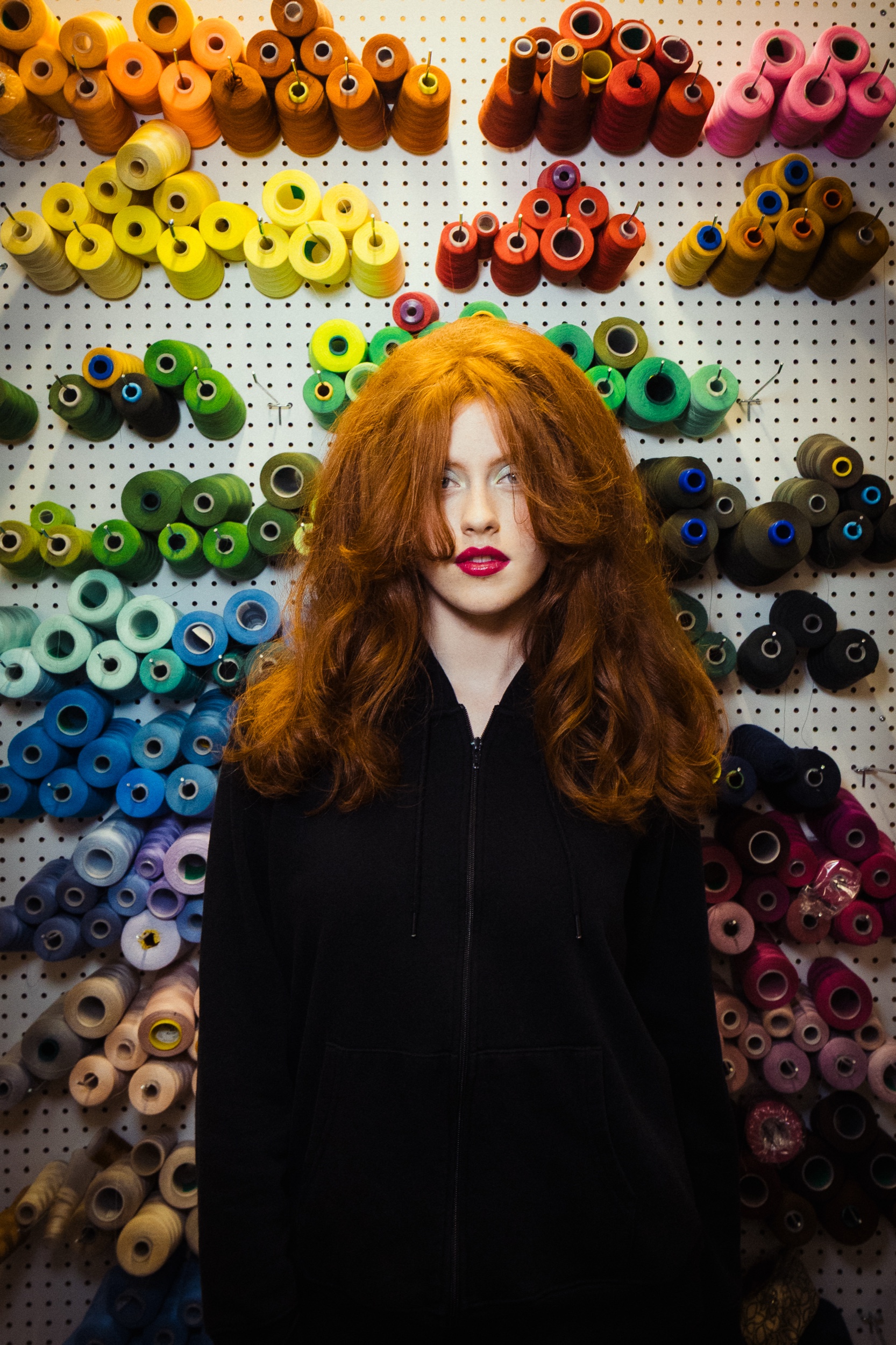 Red-haired model in an Eliza Faulkner zip jacket before a wall of thread spools in her studio, Montreal Fashion Week 2025