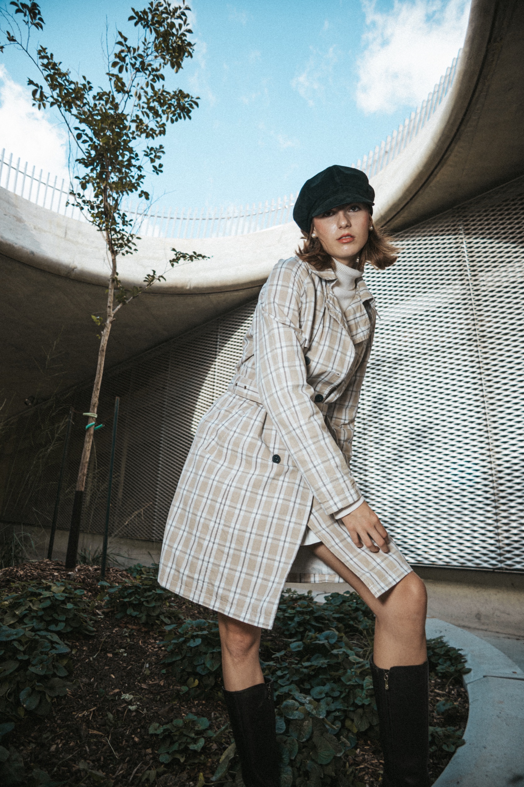 Model Arabel in a plaid coat and black cap in a curved concrete courtyard with a young tree, Montreal Fashion Week 2025