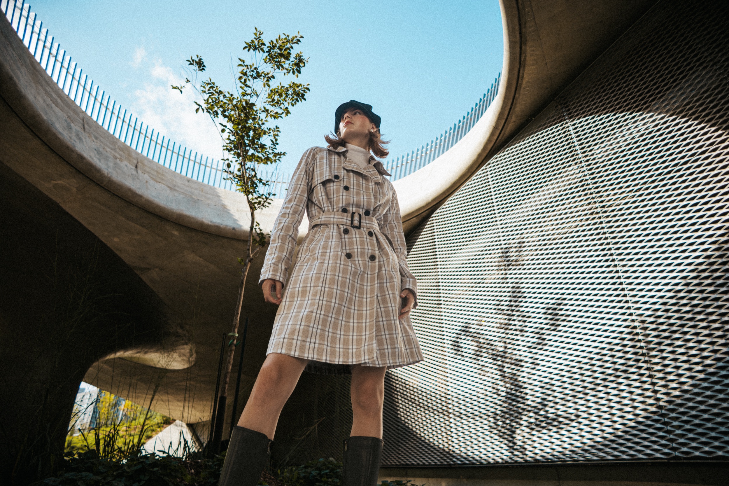 Low-angle view of model Arabel in a plaid trench coat under open sky and curved concrete, Montreal Fashion Week 2025