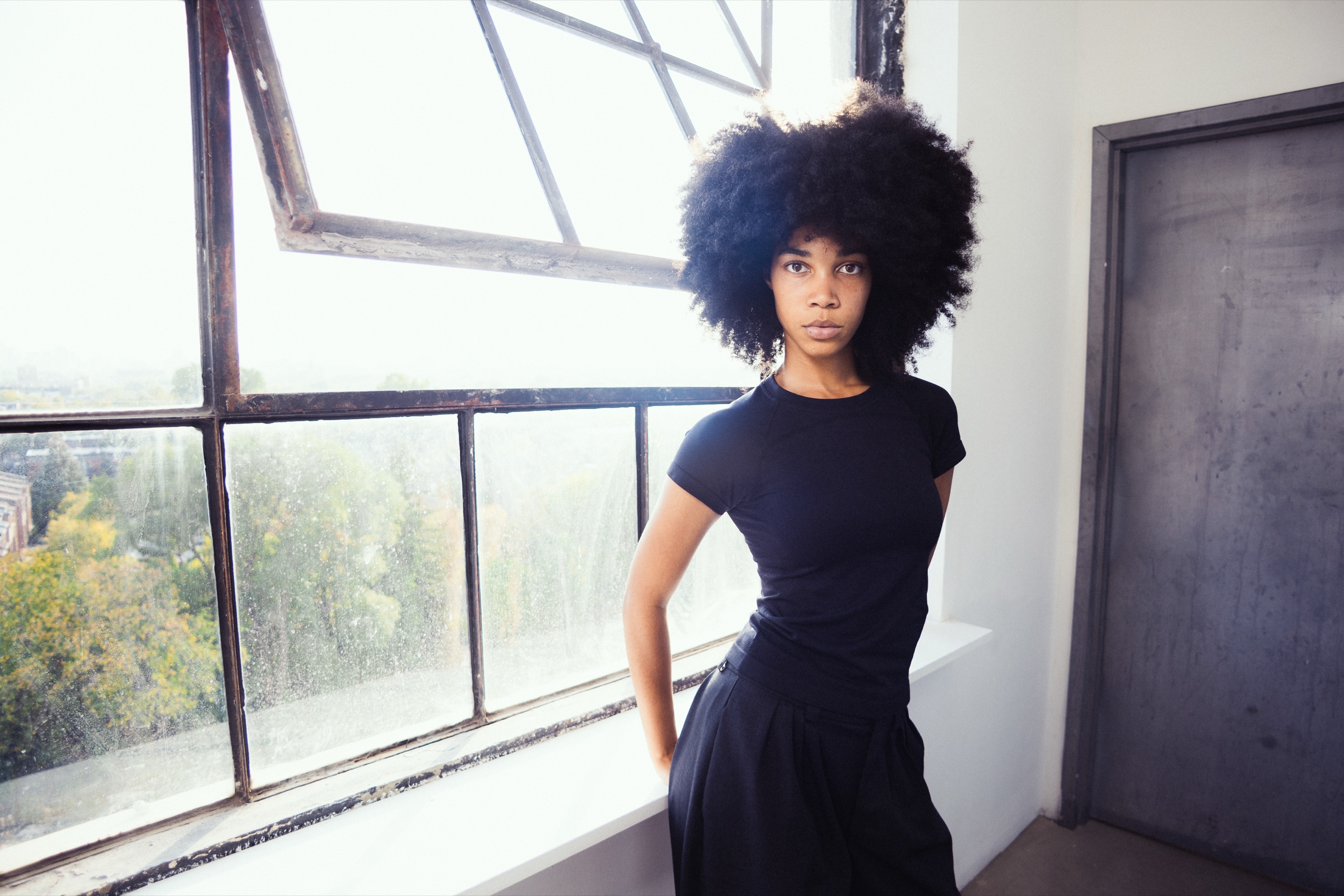 Model in a black top with voluminous natural hair stands by a tall industrial window, Montreal Fashion Week 2025