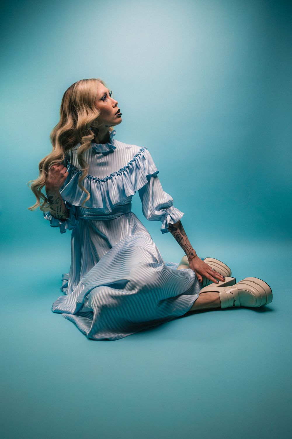 Tattooed blonde model sits in a blue striped ruffled dress and cream platforms on teal, Pastel Goths series, Edmonton