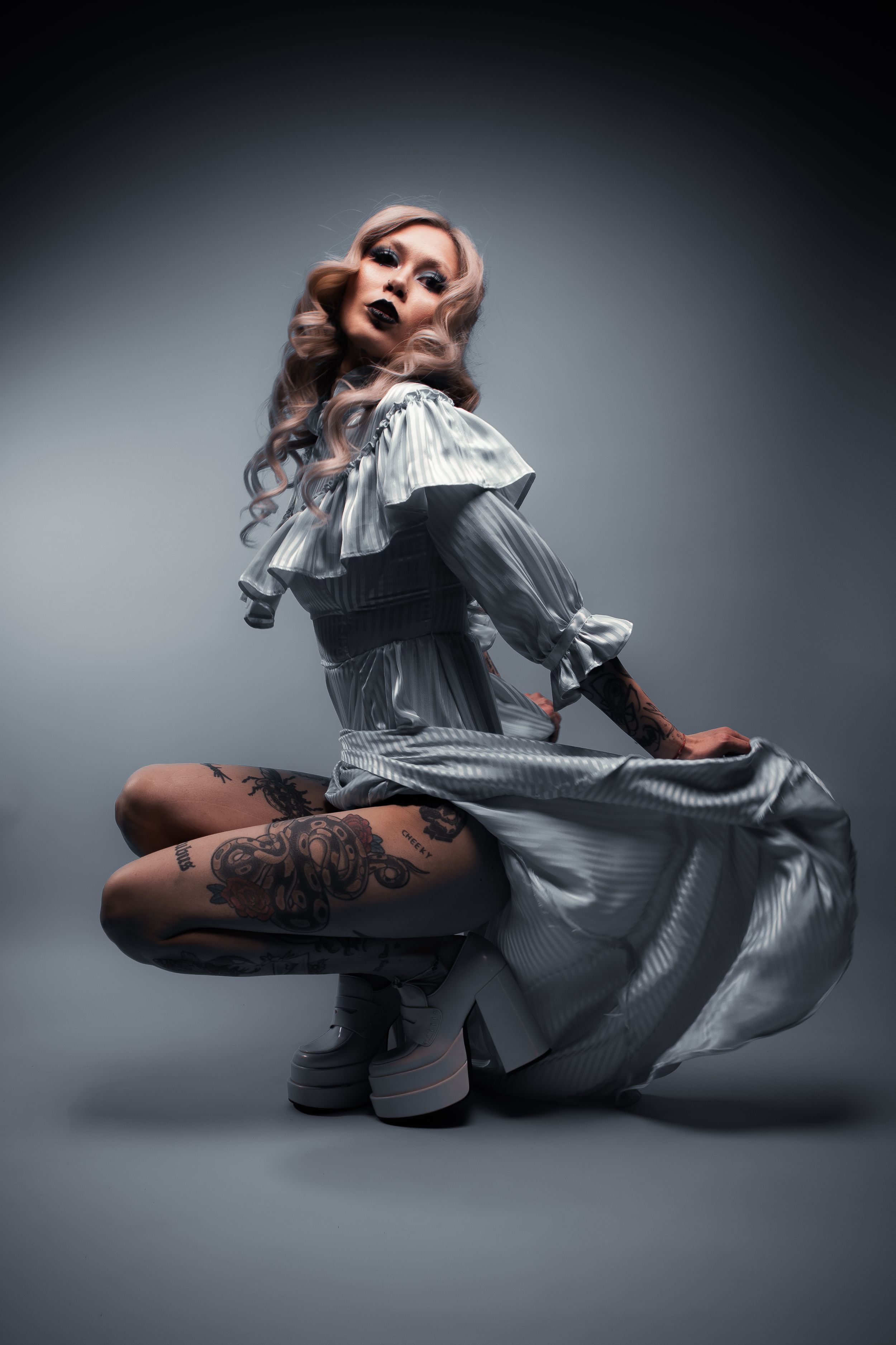 Model with platinum waves and tattoos crouches in a pale grey ruffled dress and platform boots, Pastel Goths series, Edmonton