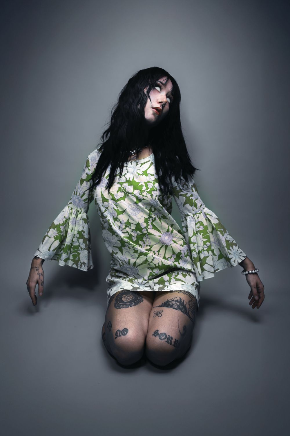 Kneeling black-haired model with tattooed knees in a green floral bell-sleeve dress on grey, Pastel Goths, Edmonton